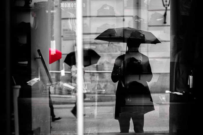 عکس خیابانی سیاه و سفید از افرادی که زیر باران قدم می زنند، یک چتر قرمز رنگ نقطه ای است.
