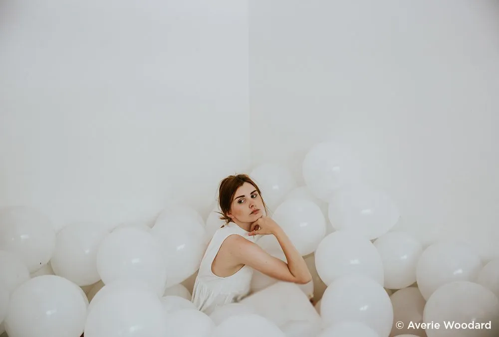 زن نشسته در گوشه اتاق میان بادکنکهای سفید در فضایی مینیمال