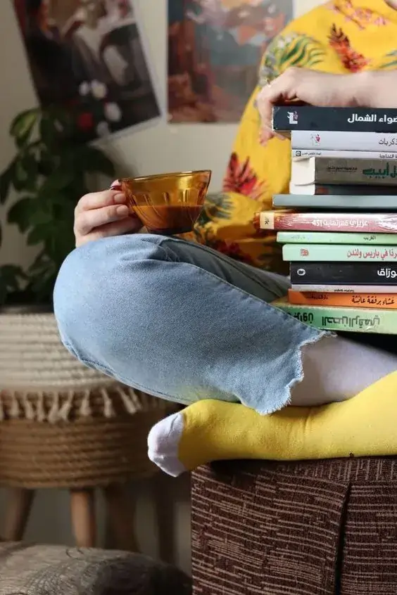 فردی نشسته با فنجان قهوه و دستهای از کتابها در خانه