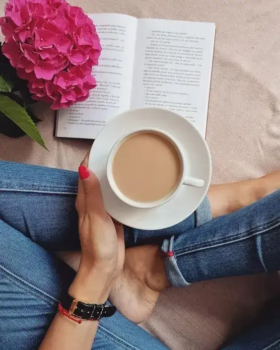 فنجان قهوه روی پای شخص در حال مطالعه کتاب با گل صورتی کنار آن