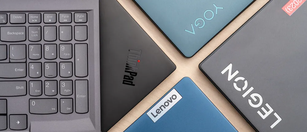 مجموعه‌ای از لپ‌تاپ‌های لنوو شامل مدل‌های ThinkPad، Yoga، Legion و IdeaPad در کنار بخشی از صفحه‌کلید خاکستری رنگ.